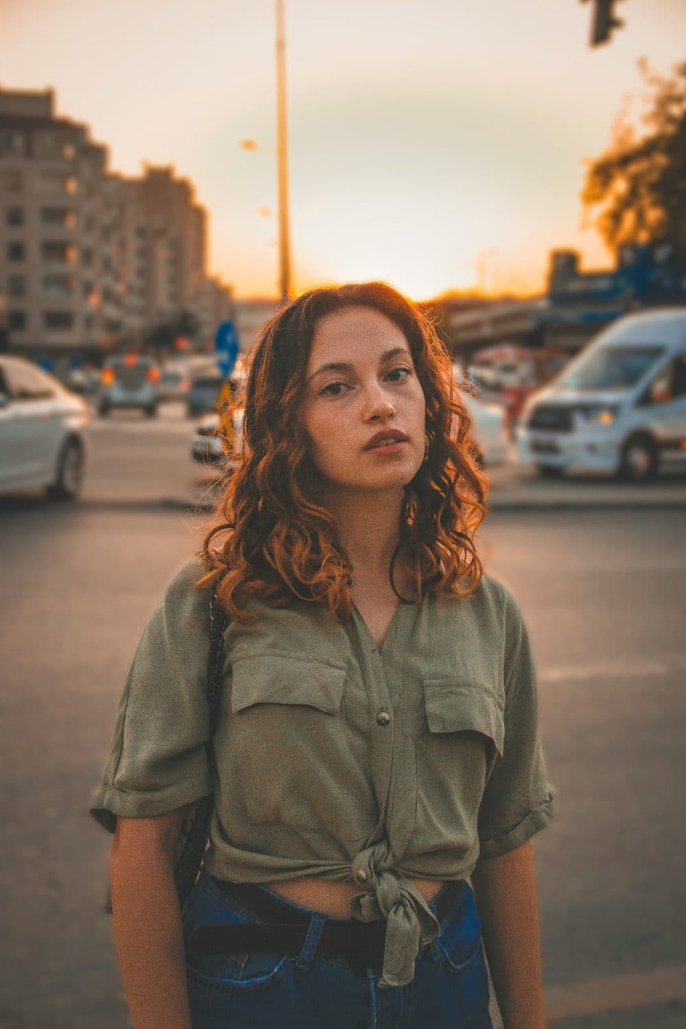 A young woman with curly auburn hair in an olive shirt standing on a city street at sunset, with blurred traffic behind her