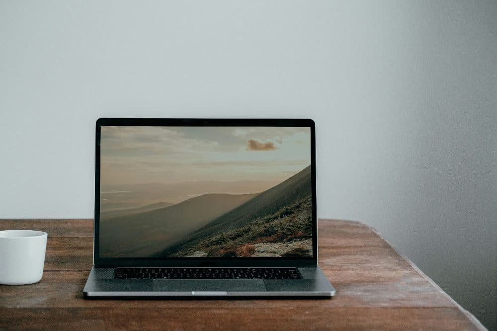 An open laptop on a wooden table showing a landscape picture full screen, with a white mug beside it