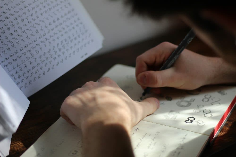 Hands inking small geometric logo marks in an open sketchbook on a wooden desk