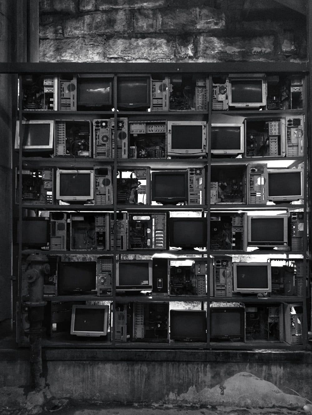 Shelves in a stone alcove stacked with old CRT monitors and open computer cases, photographed in black and white