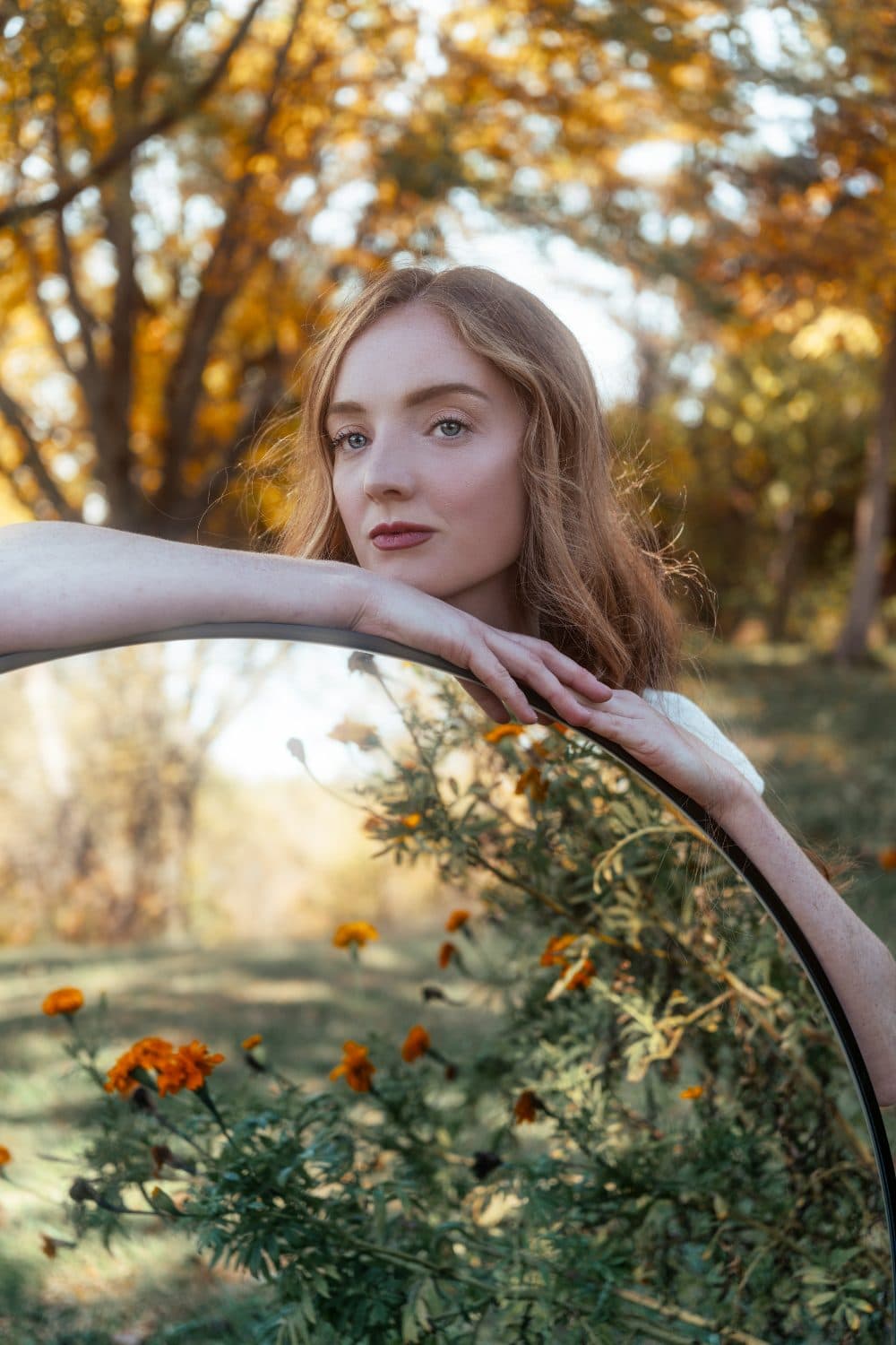 A woman with auburn hair resting her forearms on the edge of a large round mirror reflecting orange marigolds