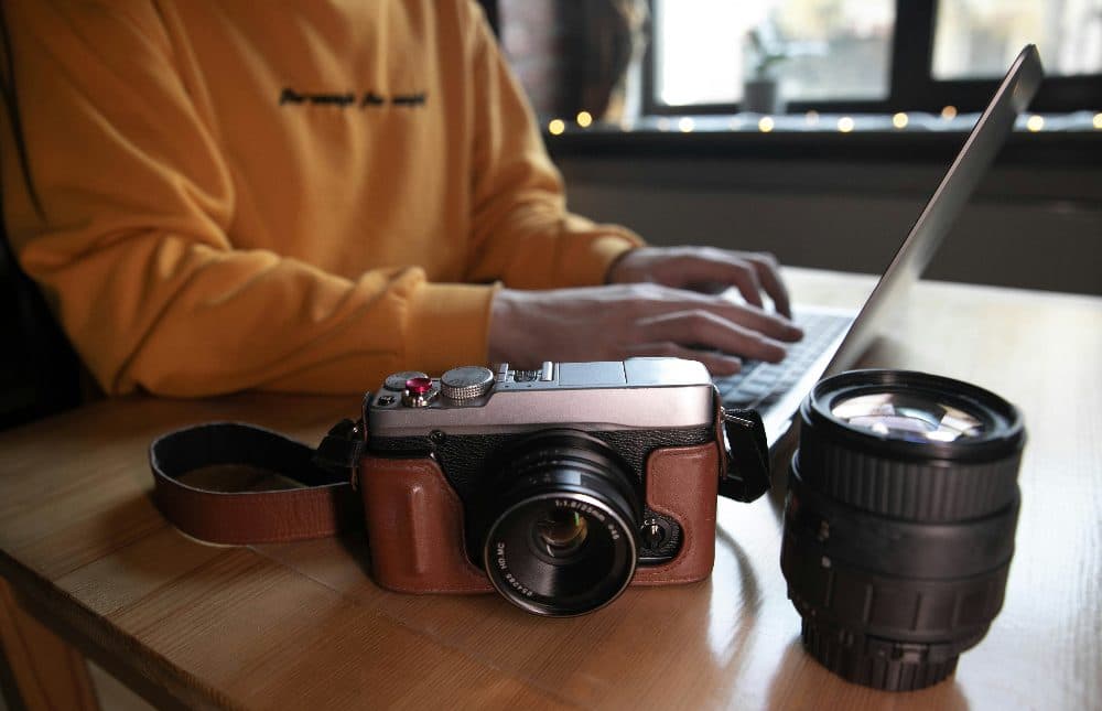 A camera and a spare lens on a wooden table while someone types on a laptop behind them