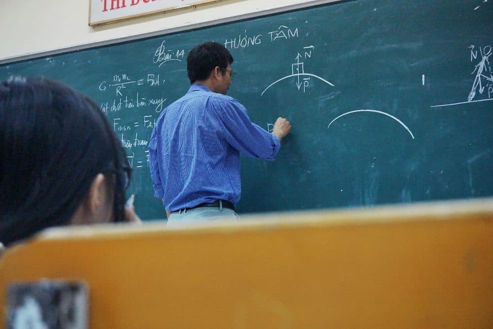 A teacher in a checked shirt writing formulas on a green chalkboard, with a student’s desk in the foreground
