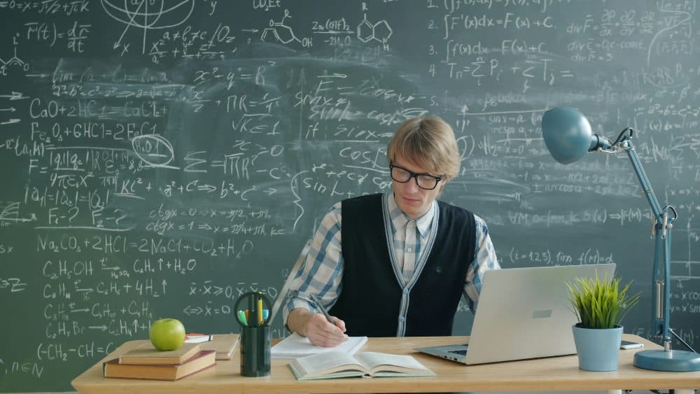 A young man in glasses writing in a notebook at a desk with a laptop, in front of a chalkboard of equations
