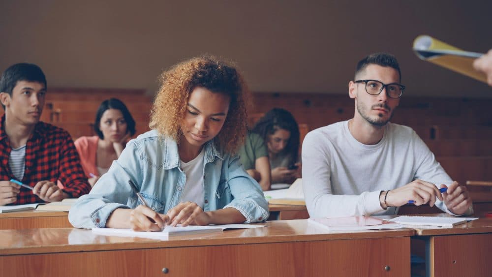 A young woman in a denim jacket writing on paper at a desk in a lecture hall, with other students around her
