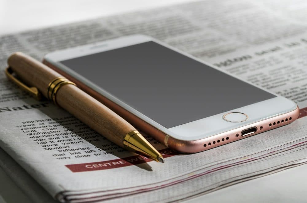 A rose gold smartphone lying on a folded newspaper next to a wooden ballpoint pen