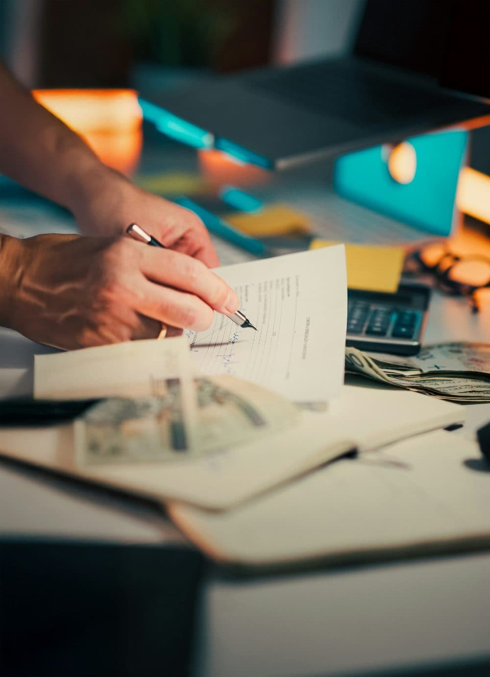 A hand holding a fountain pen over a printed paper form on a desk, next to a calculator, a laptop and some banknotes