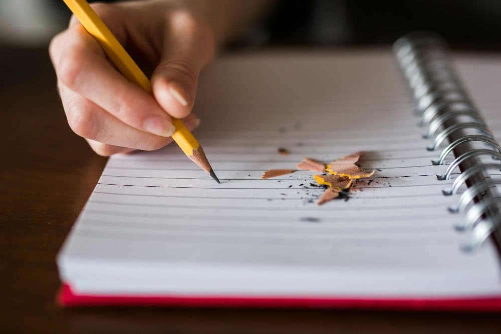 A hand holding a yellow pencil over a lined spiral notebook with pencil shavings scattered on the page