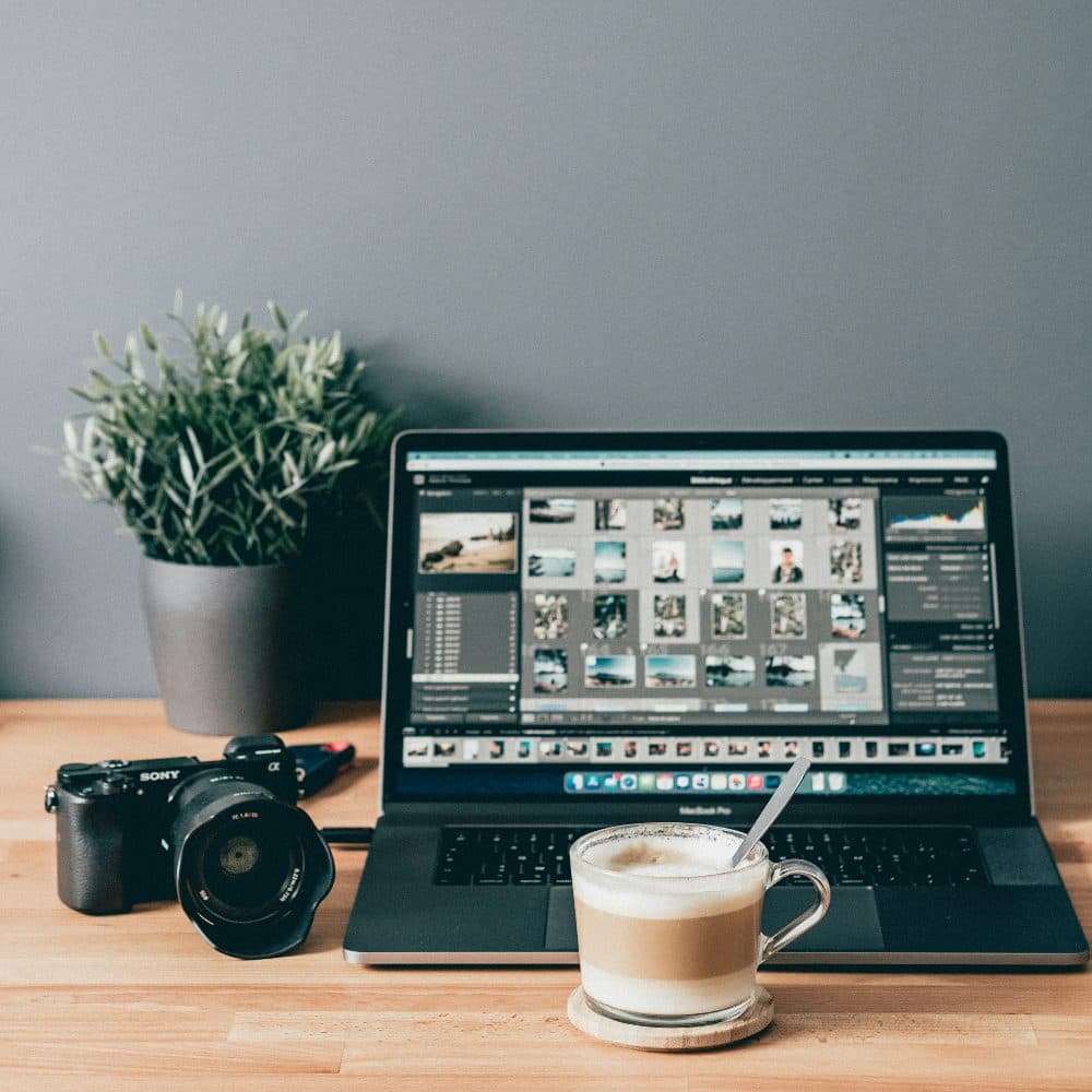 A laptop showing a photo library beside a camera, a lens and a cup of coffee on a wooden desk