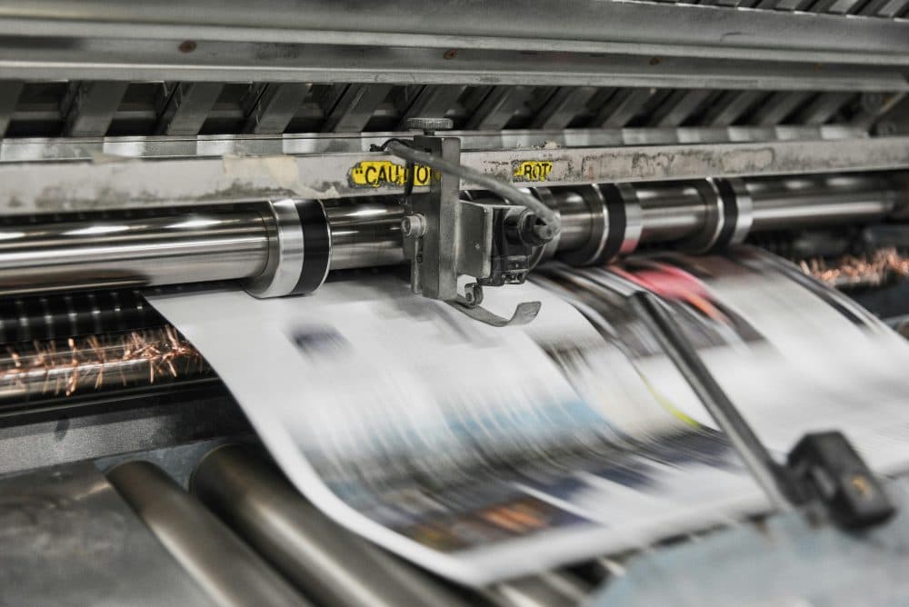 A industrial printing press feeding a freshly printed, motion-blurred sheet of paper past steel rollers