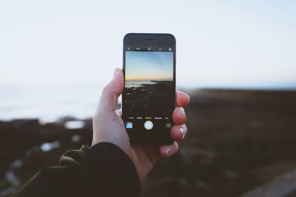 A hand holding a smartphone with its camera app open, framing a sunset over a rocky shoreline