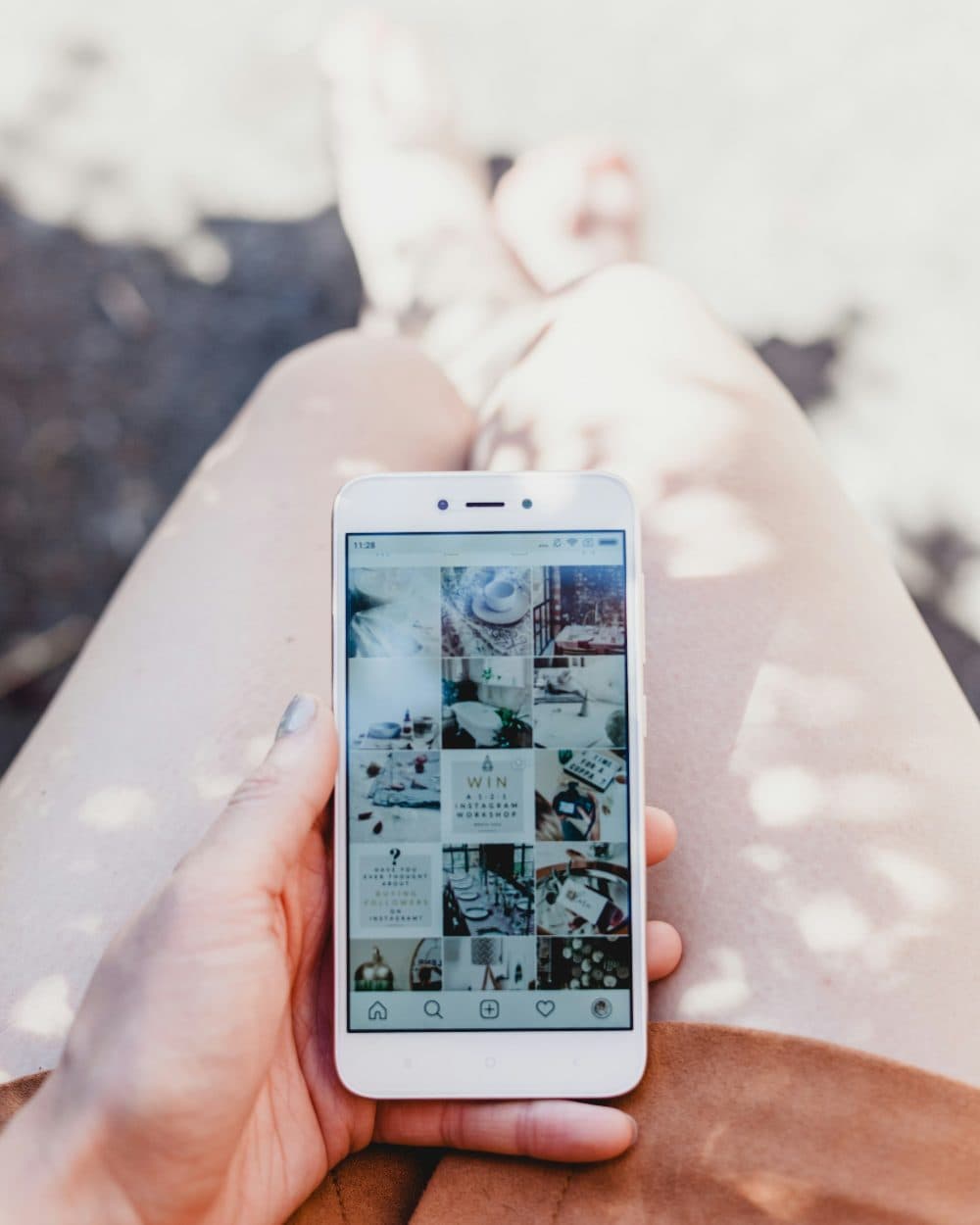 A hand holding a white smartphone that shows a grid of square photo thumbnails, held above a person’s lap