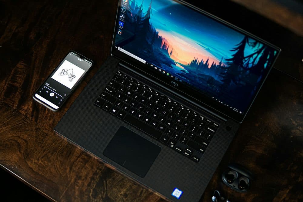 A black laptop showing a Windows desktop with a forest sunset wallpaper, on a dark desk beside a phone and earbuds