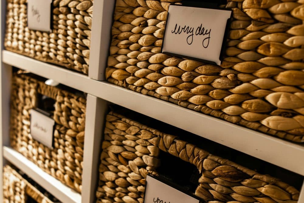 Woven wicker storage baskets in a white shelving unit, each fitted with a small handwritten label card