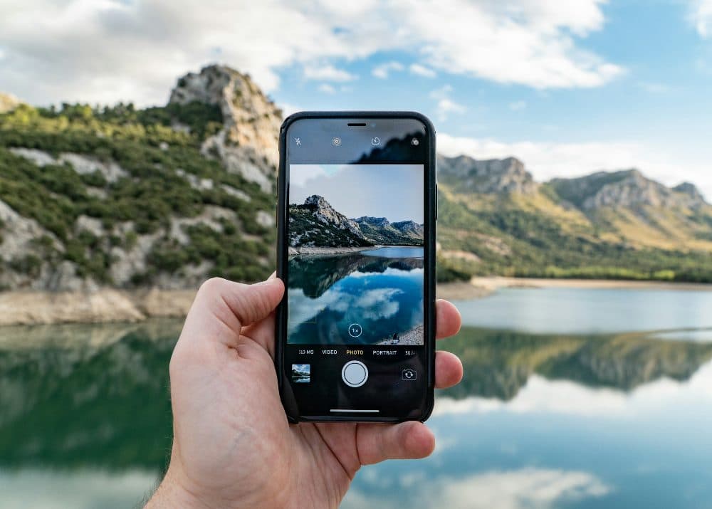 A hand holding up a phone to photograph a mountain lake, the same view on its screen