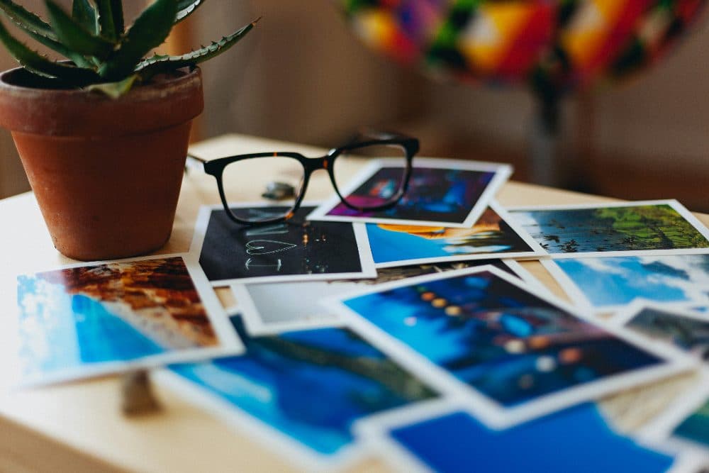 Printed photographs spread out on a table beside a pair of glasses and a plant pot