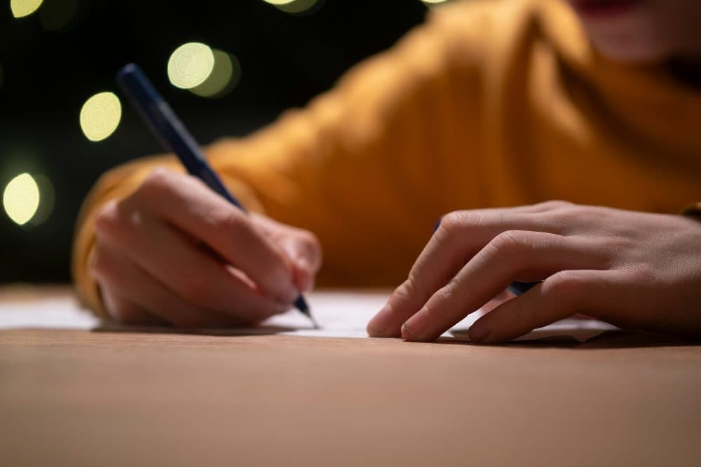 A hand holding a blue pen and writing on a sheet of paper on a wooden desk, with the other hand resting beside it