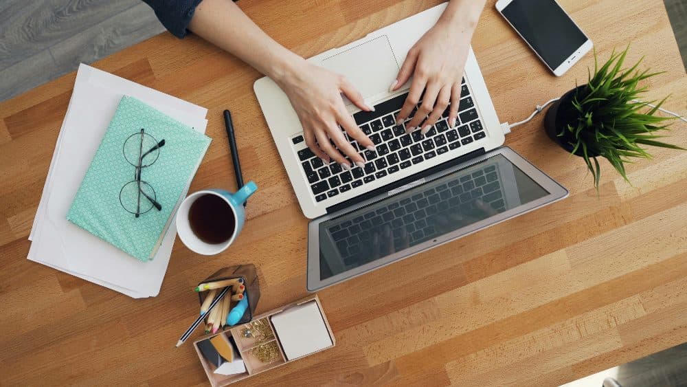 Top-down view of hands typing on a laptop at a wooden desk with a notebook, tea and a plant