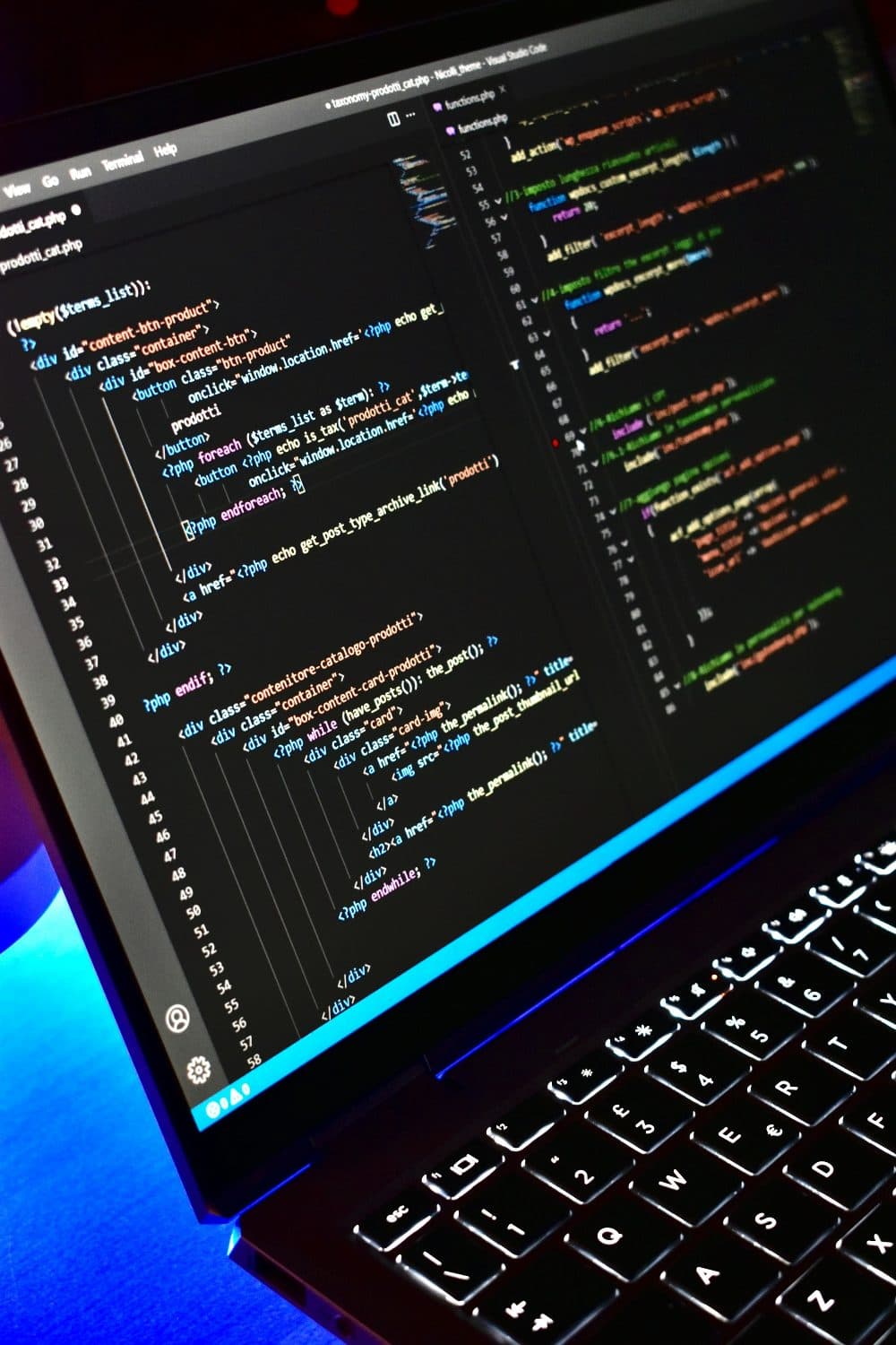 A laptop on a desk in blue light, its screen filled with lines of code in a dark editor, backlit keyboard in front