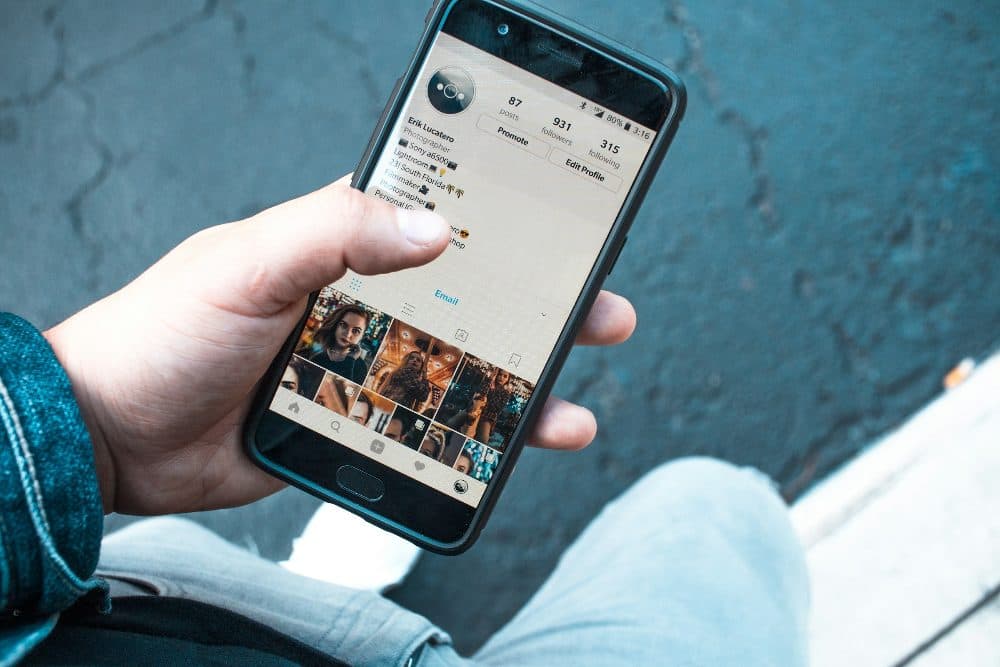 A hand holding a smartphone that shows a profile page with a round profile picture and a grid of photos