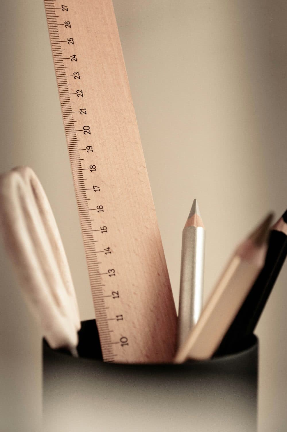 A wooden ruler with centimetre markings standing in a black pot beside a silver pencil and other stationery