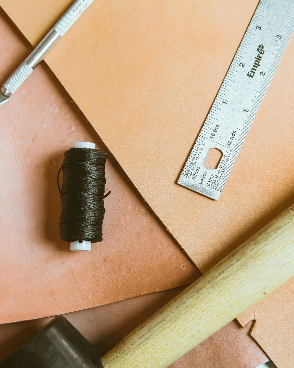 A steel ruler and a spool of dark thread lying on sheets of tan leather on a workbench