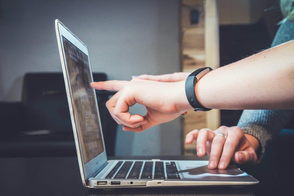 A hand wearing a fitness band pointing at the screen of an open laptop while another hand rests on the trackpad