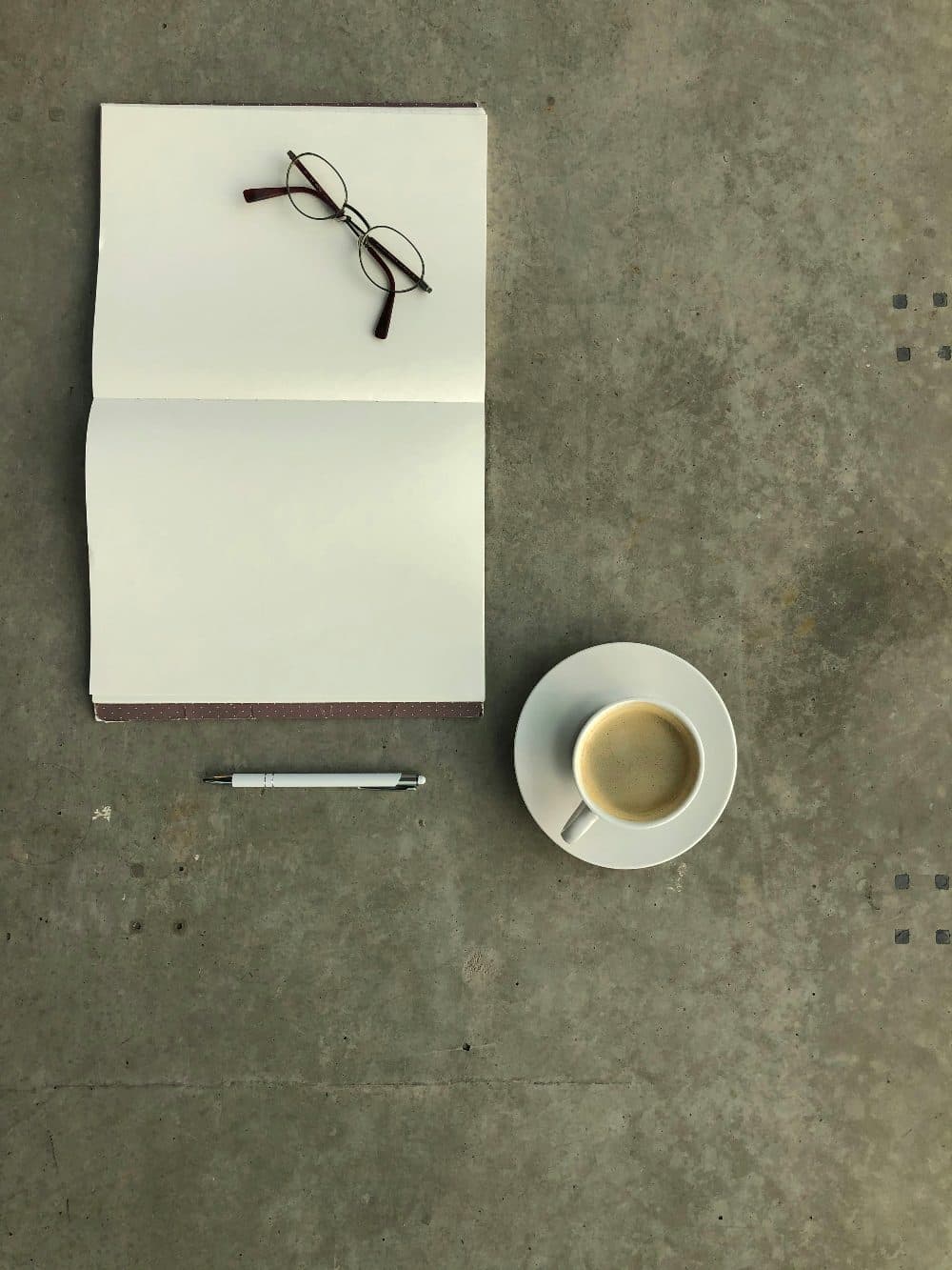 An open blank notebook with glasses, a pen and a cup of coffee on a grey concrete surface