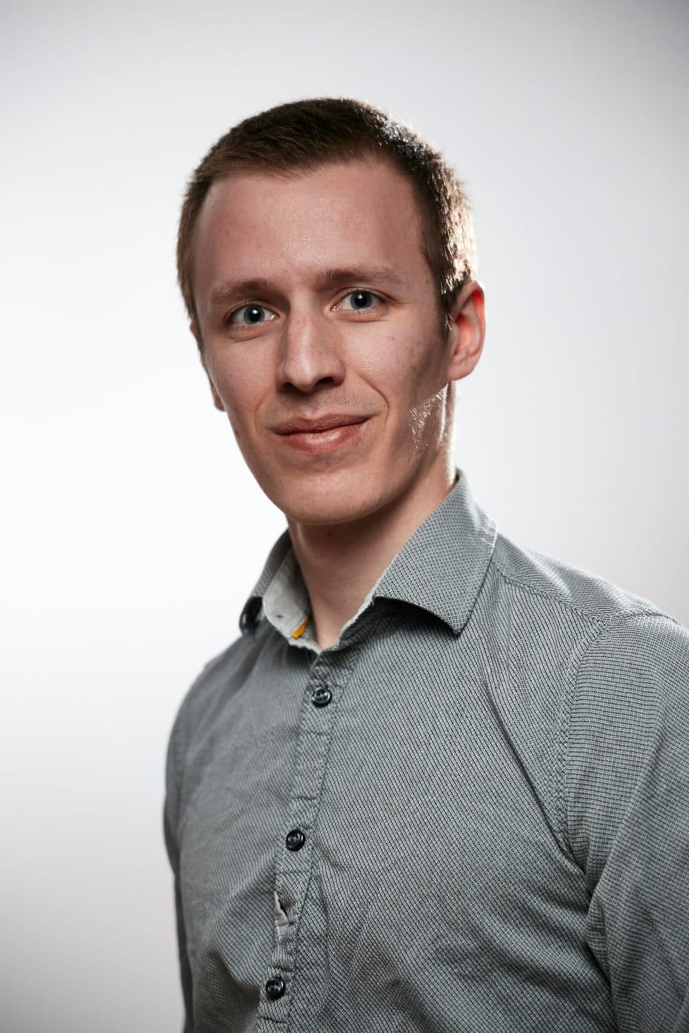 A smiling man in a grey button-up shirt photographed head and shoulders against a white wall