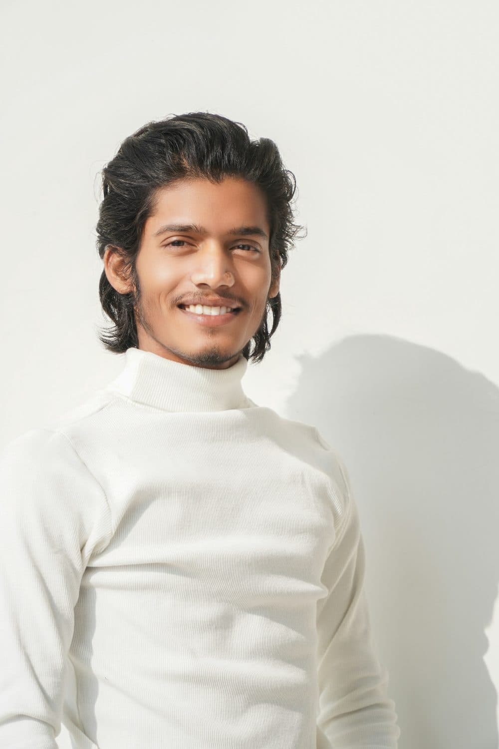 A smiling young man with swept-back dark hair in a white polo-neck jumper, standing against a plain light wall