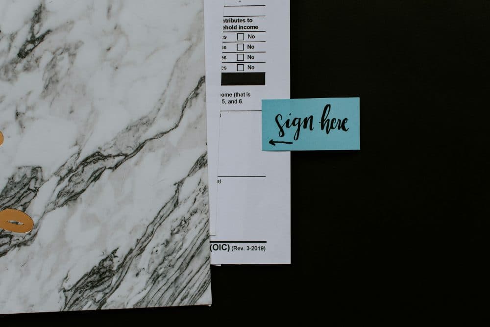 A blue “sign here” sticky note with an arrow placed beside a printed form on a marble-patterned folder