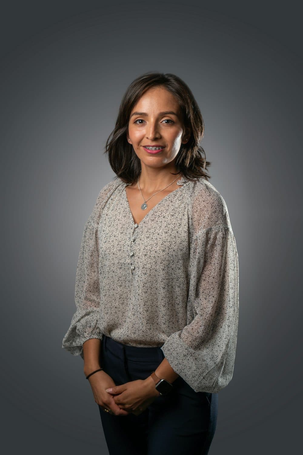A woman in a light patterned blouse photographed in a studio against a grey backdrop