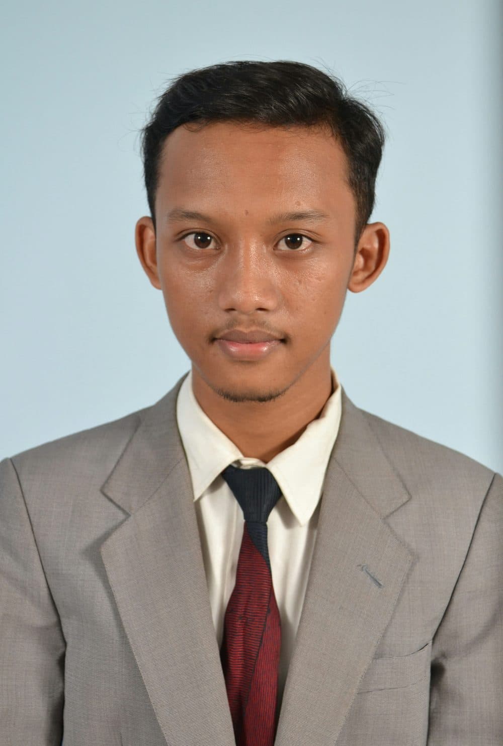 A young man in a grey suit and dark tie facing the camera against a pale blue background