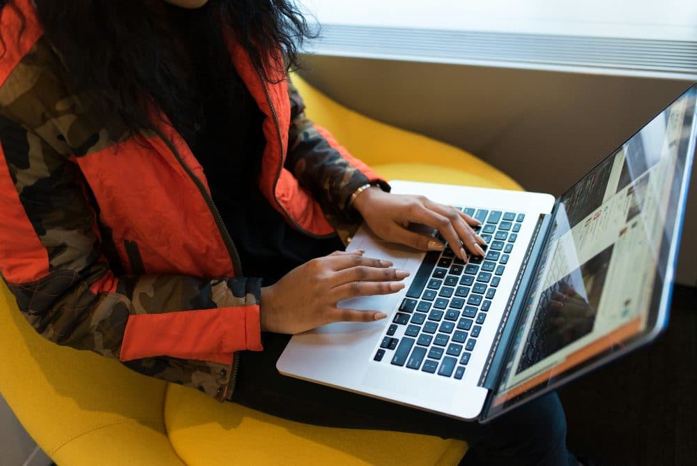 A woman in a red and black jacket typing on a laptop while sitting in a yellow chair