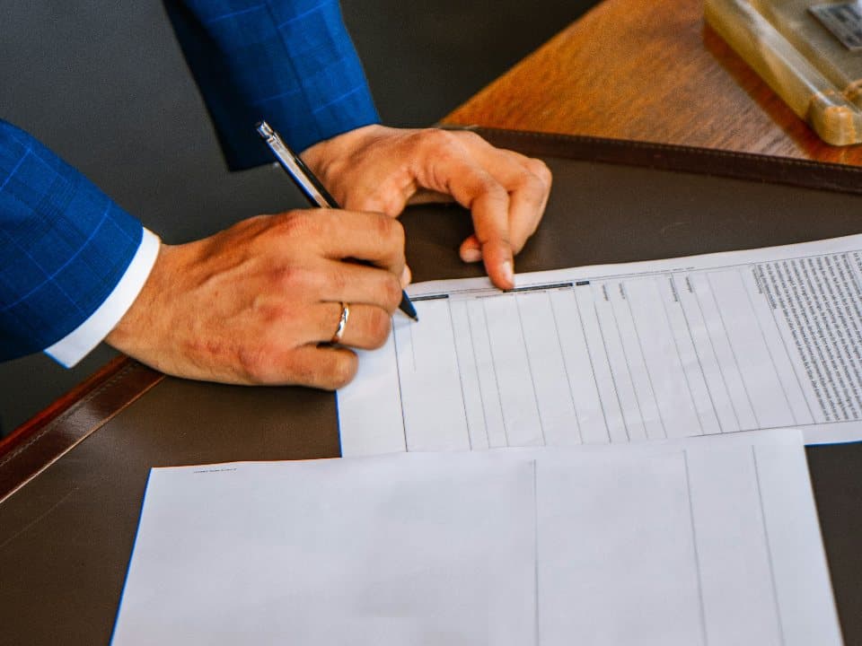 A hand in a blue suit sleeve signing a printed form with a pen on a wooden desk