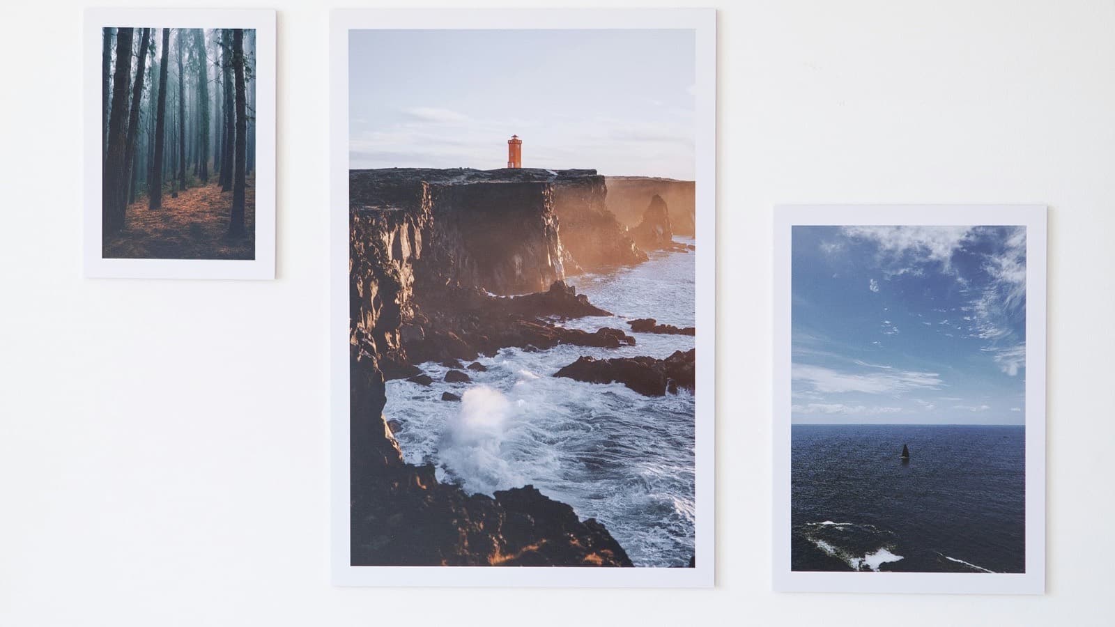 Three photographic prints of a misty forest, a lighthouse on a sea cliff and a small boat hung on a plain white wall