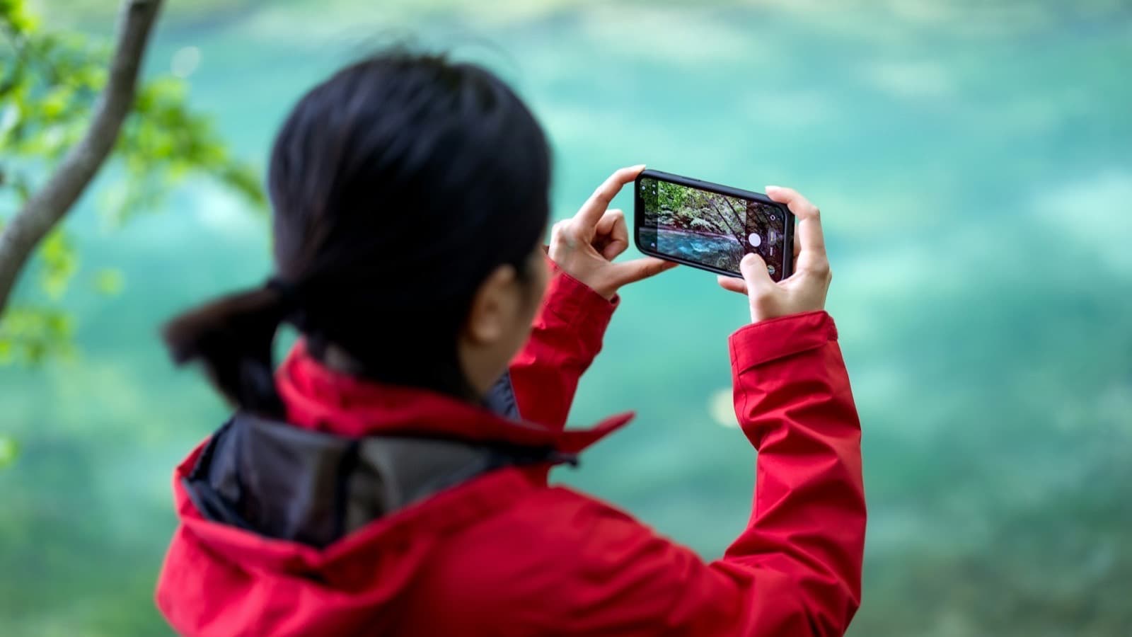A person in a red jacket, seen from behind, holding a smartphone sideways to photograph a turquoise lake