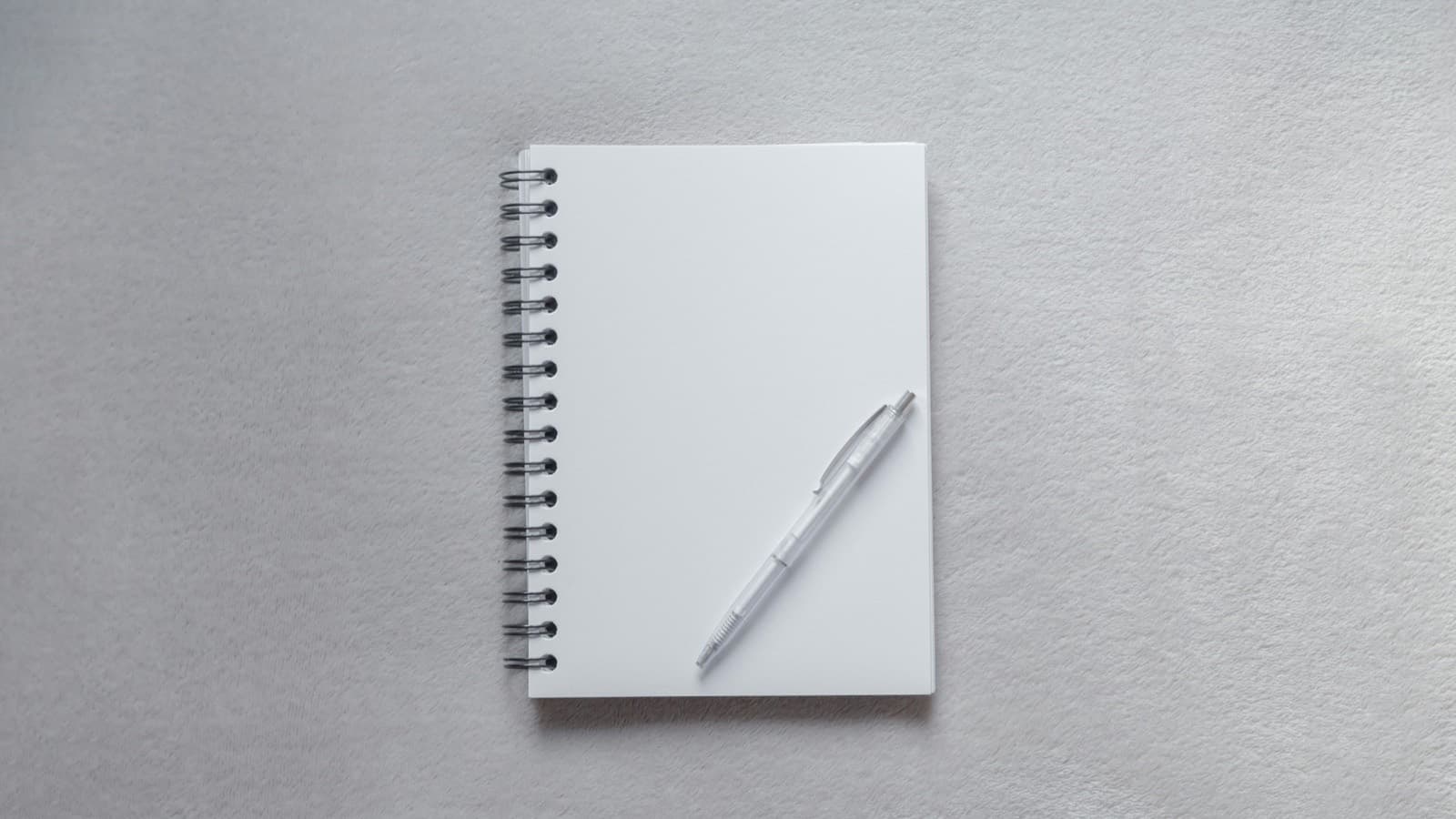 A clear plastic ballpoint pen lying on a blank white spiral-bound notebook on a grey fabric surface, seen from above