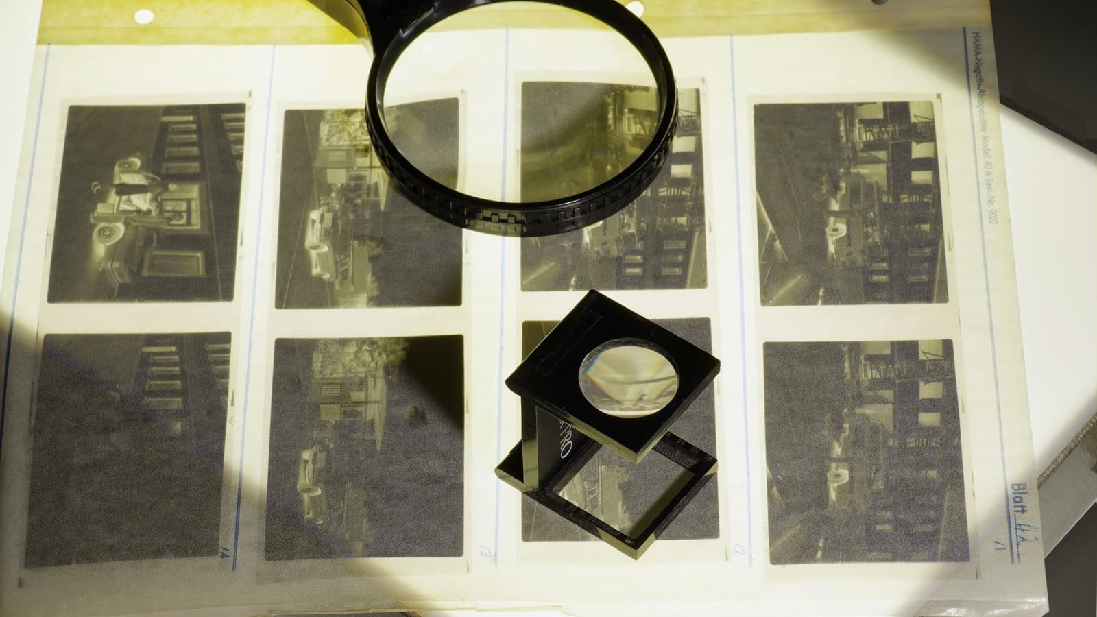 A black magnifying glass and a small folding loupe resting on a sheet of black-and-white film negatives on a light table