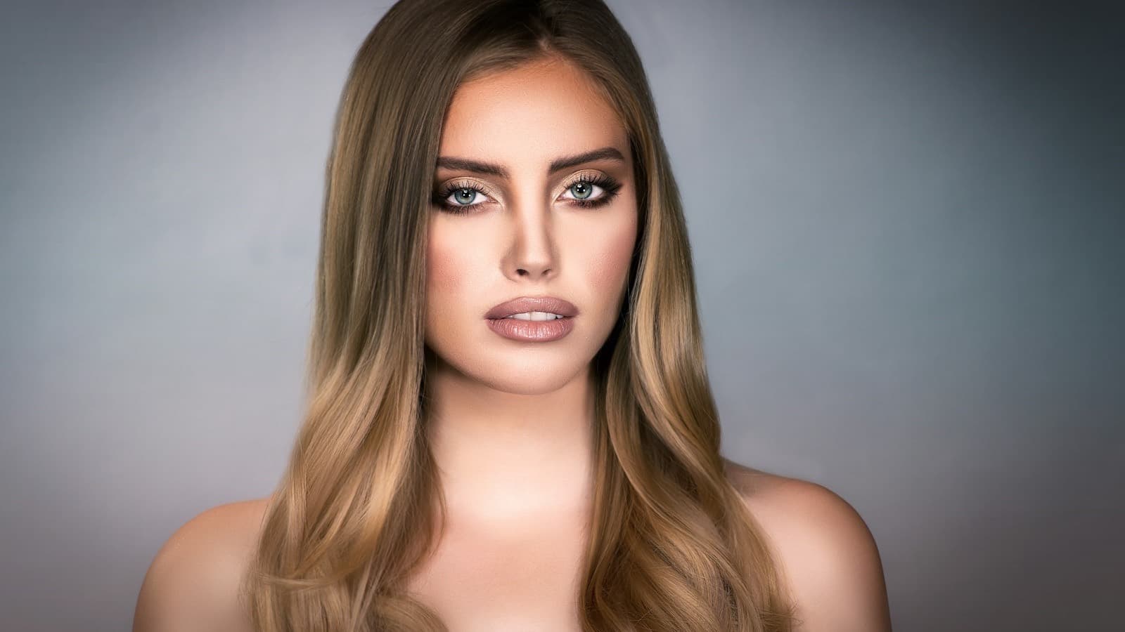 A young woman with long light brown hair looking straight at the camera against a plain grey studio background