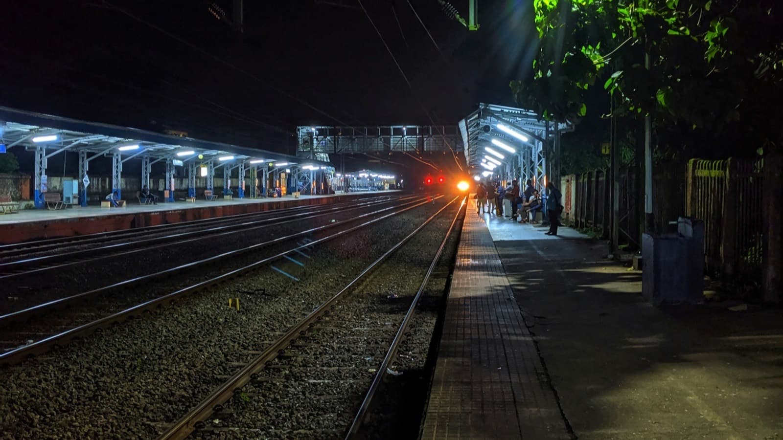 A train headlight glowing at the far end of a railway station platform at night, with lit platform lamps