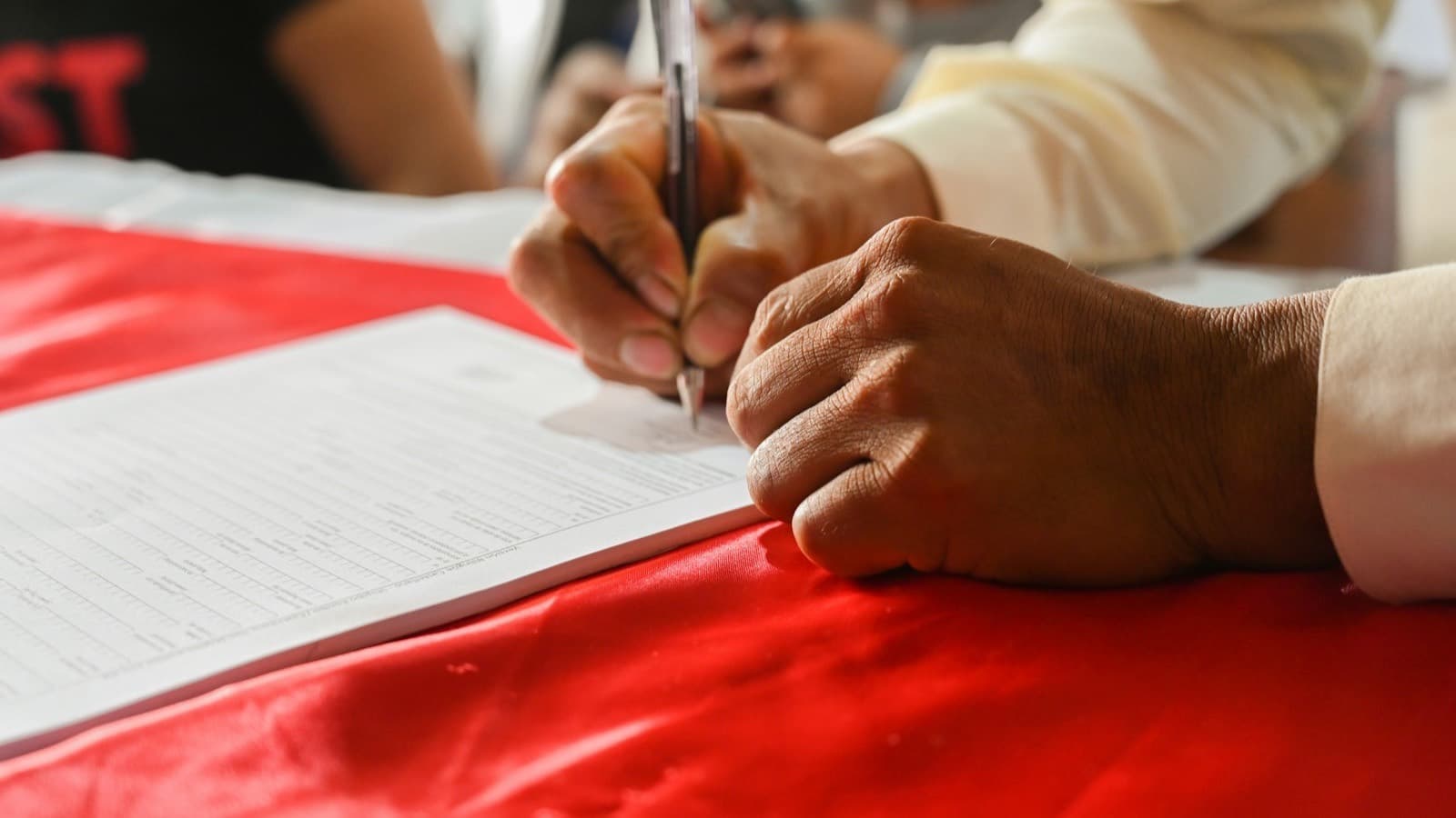 A hand holding a ballpoint pen over a printed paper form lying on a table covered with red satin cloth