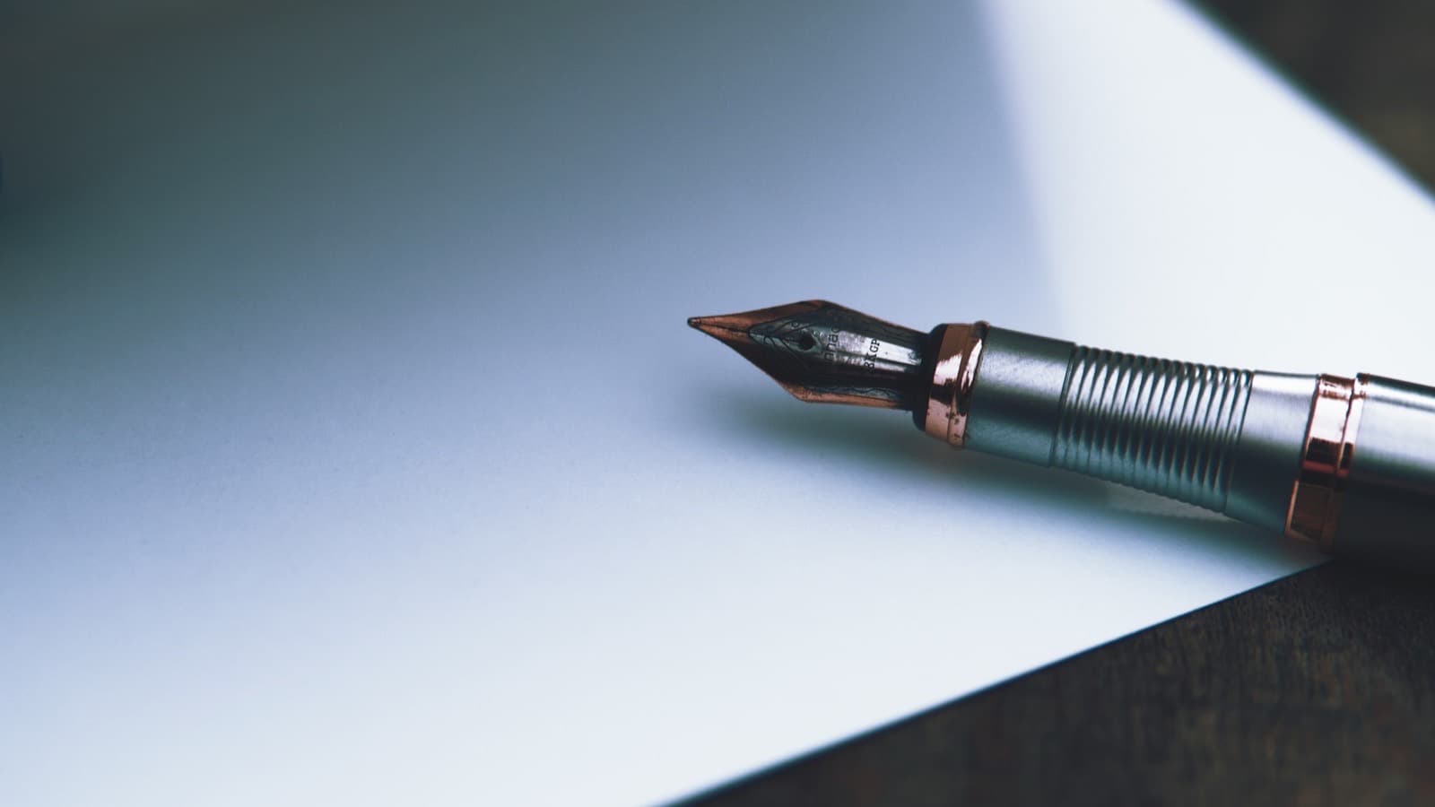 A silver and copper fountain pen lying on a blank white sheet of paper on a dark wooden desk